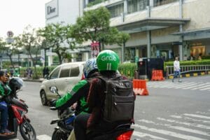 A Grab motorcycle taxi carrying a passenger on a busy city street, with the rider wearing a green Grab helmet. The background features a shopping mall and other motorbike riders. Is Grab a Good Stock to Buy?